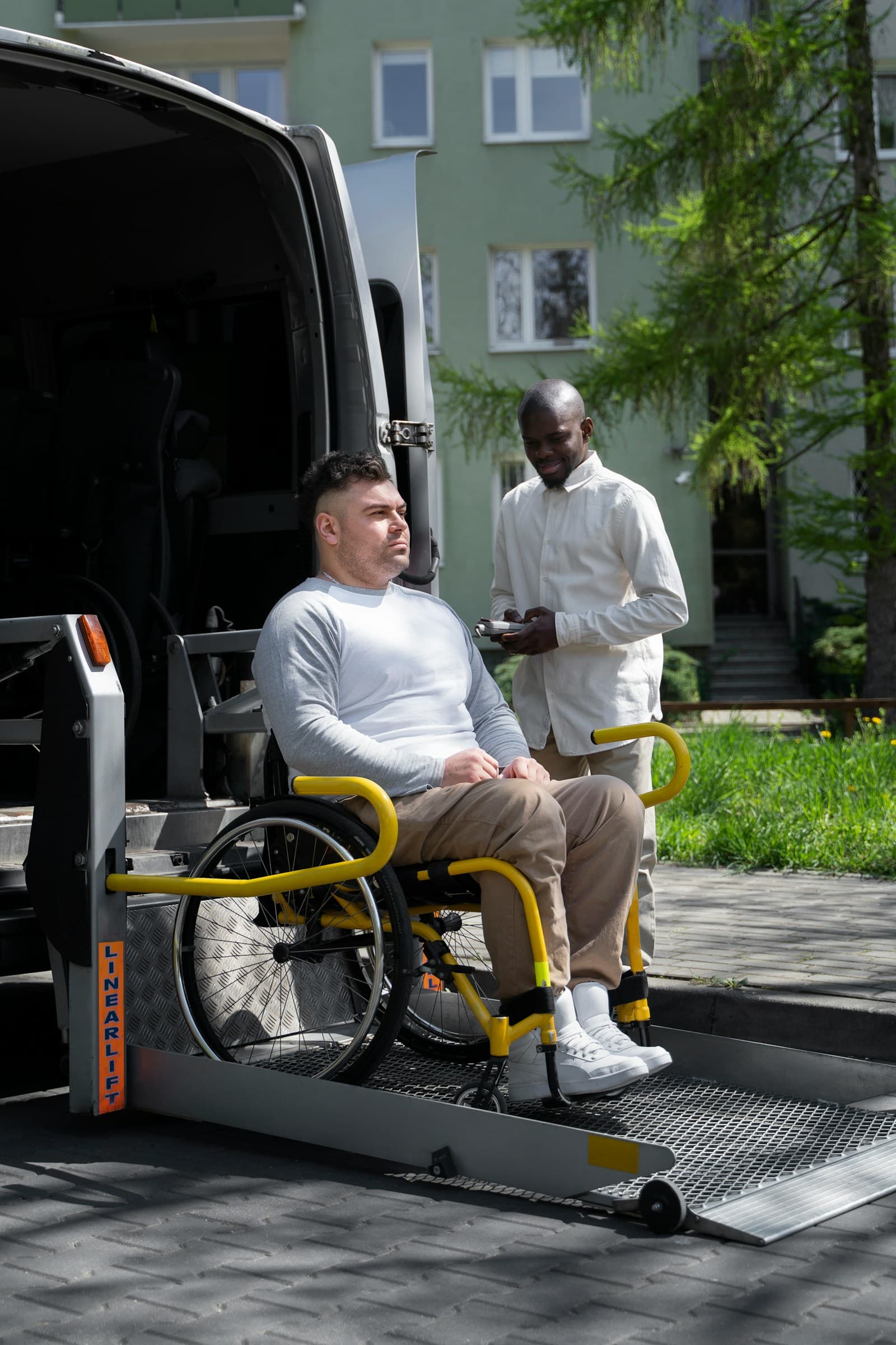 A man sitting in a wheelchair next to an accessible van, being assisted by a caregiver
